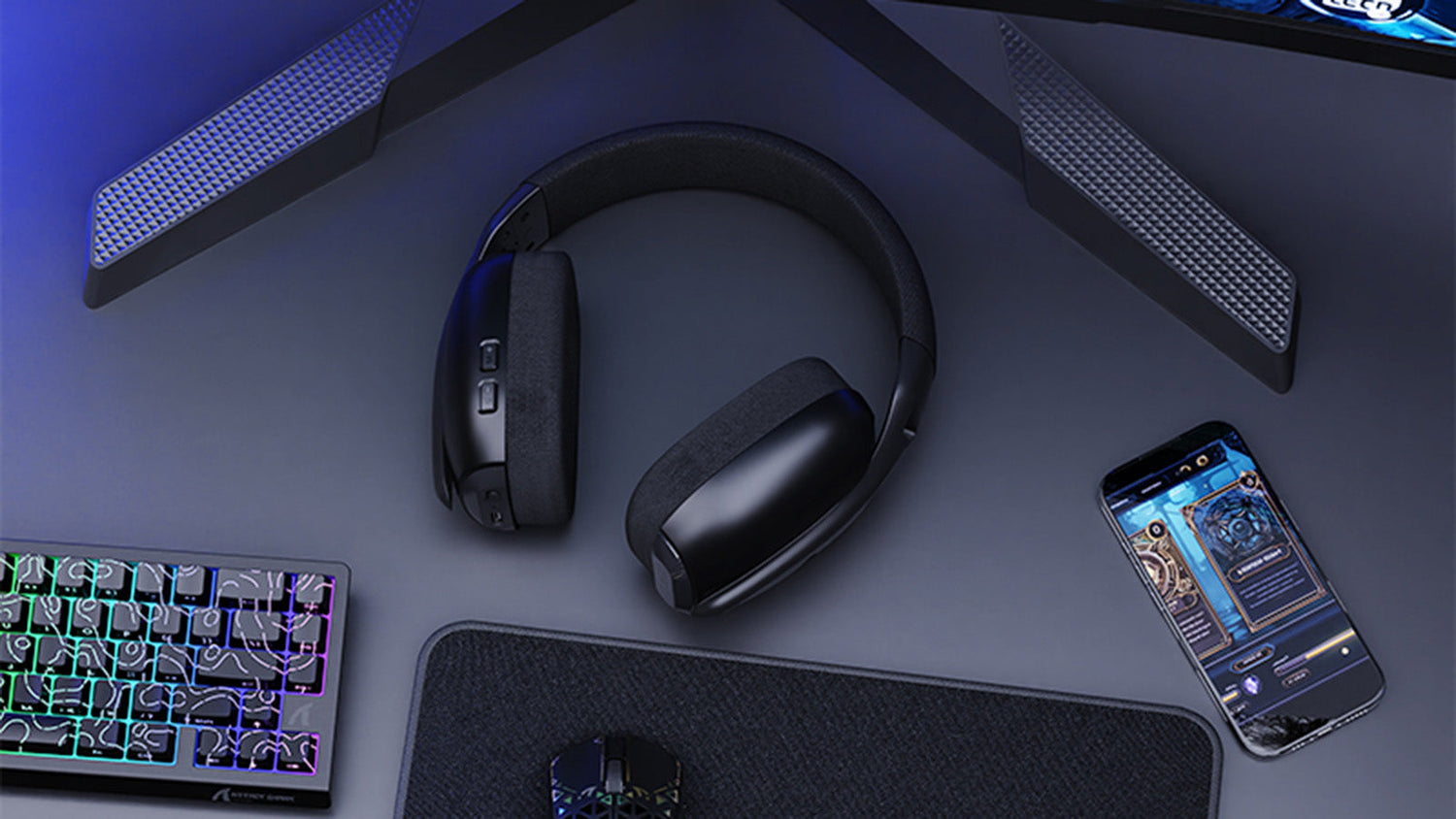 Black wireless gaming headphones on a dark desk with RGB keyboard and smartphone displaying a gaming interface. The setup is illuminated with blue ambient lighting, creating a modern gaming atmosphere